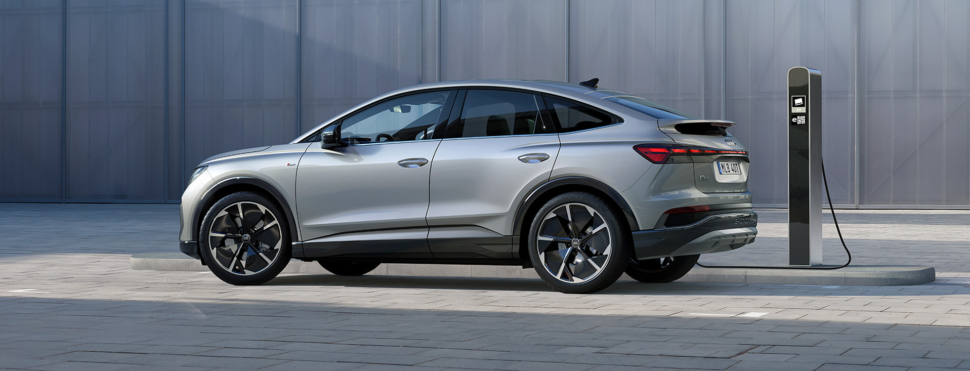 Audi Q4 Sportback e-tron, i färgen silvergår metallic, står parkerad framför en betongvägg, med laddaren i.