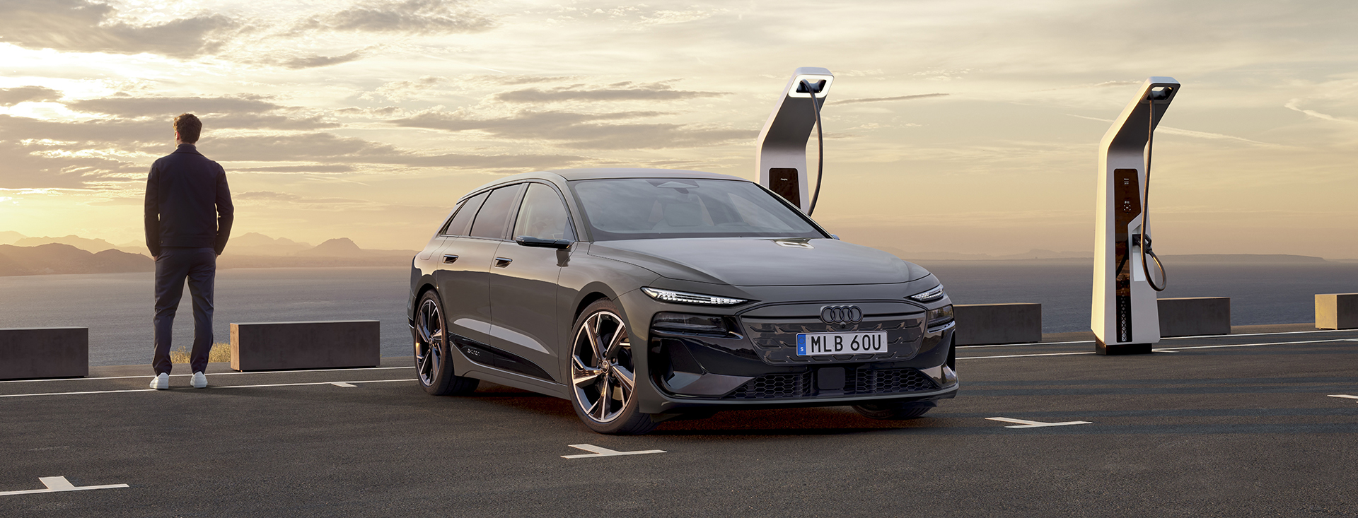 Nya A6 Avant e-tron står parkerad vid en laddstolpe vid havet. Man står och blickar mot horisonten i solnedgången