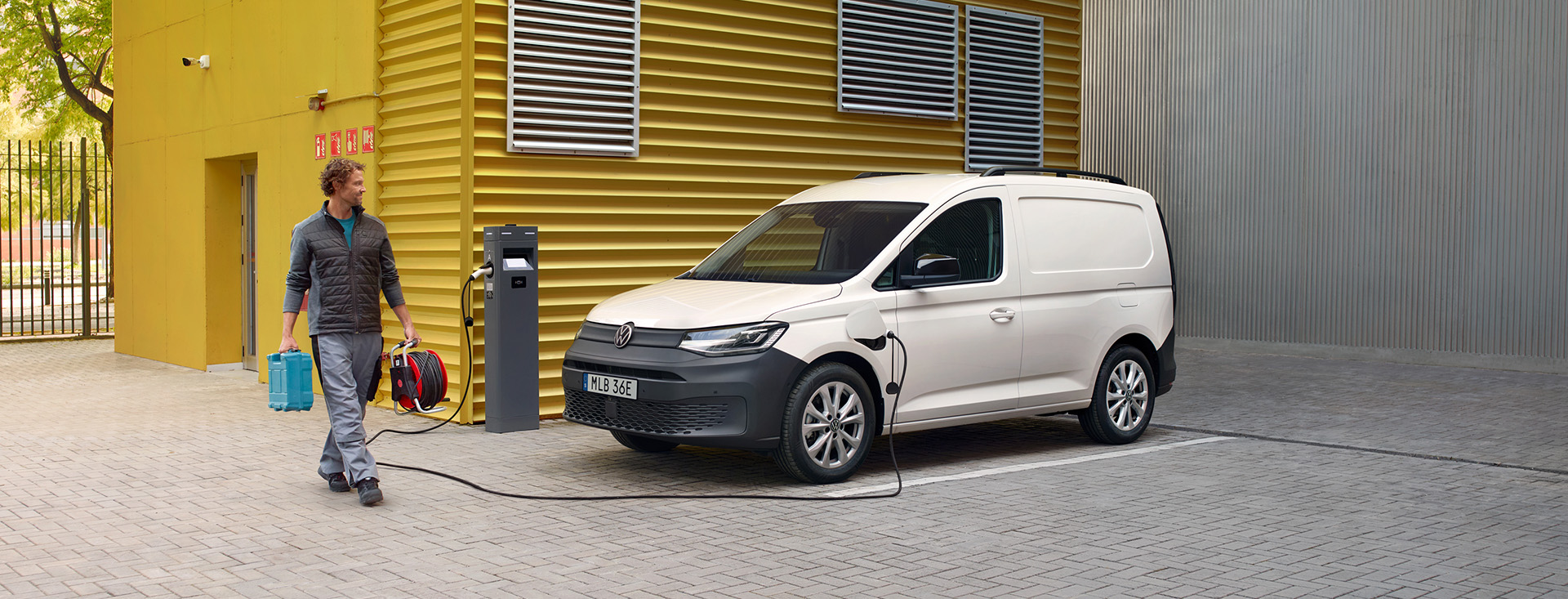 En man som bär på kabelvinda och verktygslåda går mot Caddy Cargo laddhybrid som står parkerad framför en gul vägg med laddsladden i.